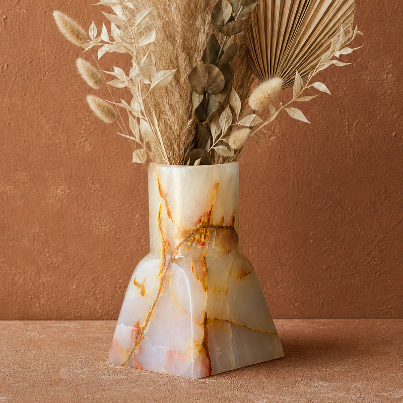 A sculptural vase made from polished onyx with natural gold and orange veining, filled with dried botanicals, set against a textured terracotta background.