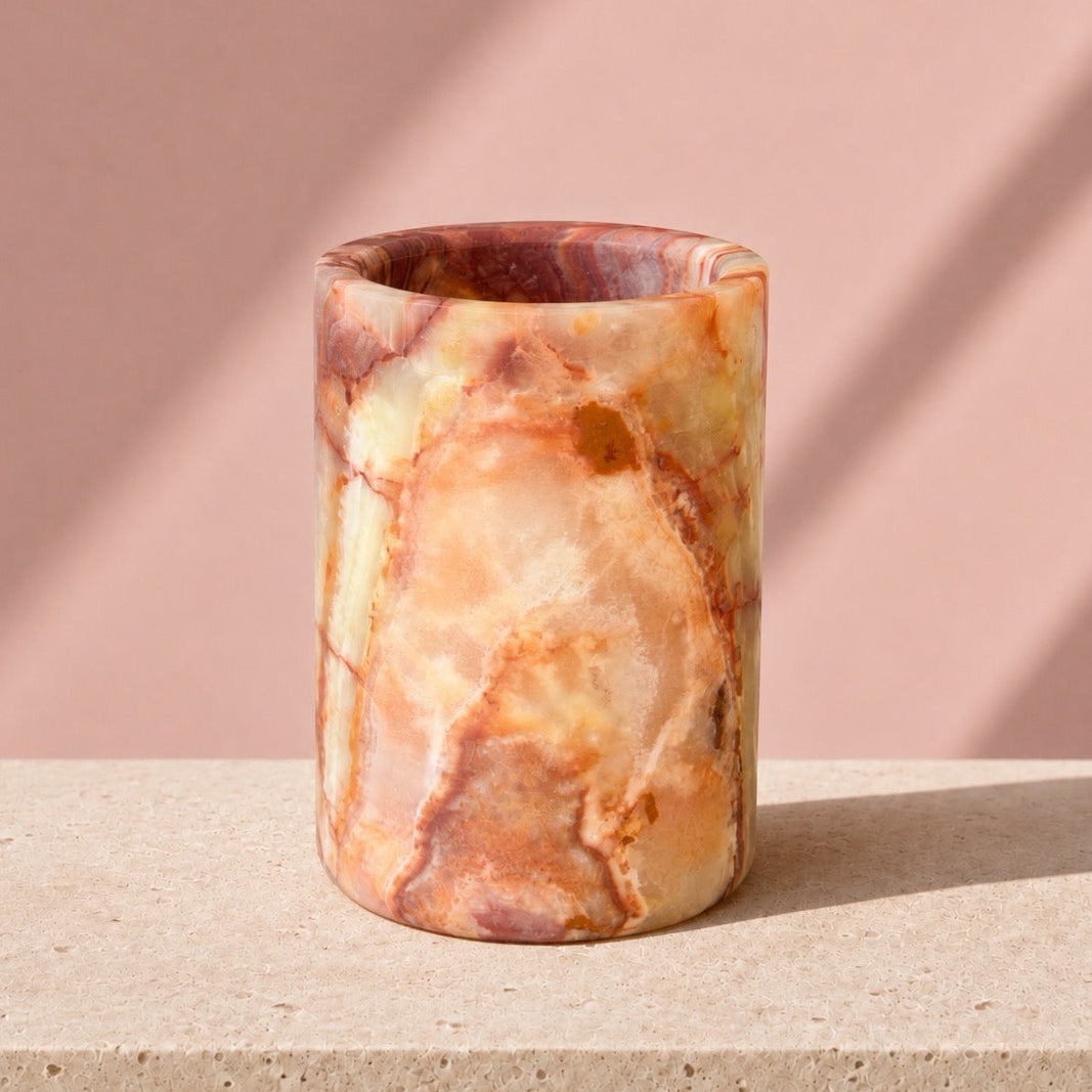 A cylindrical onyx stone vase with a marbled pattern of beige, orange, and reddish-brown, standing on a light stone surface against a dusty pink background with soft diagonal shadows.