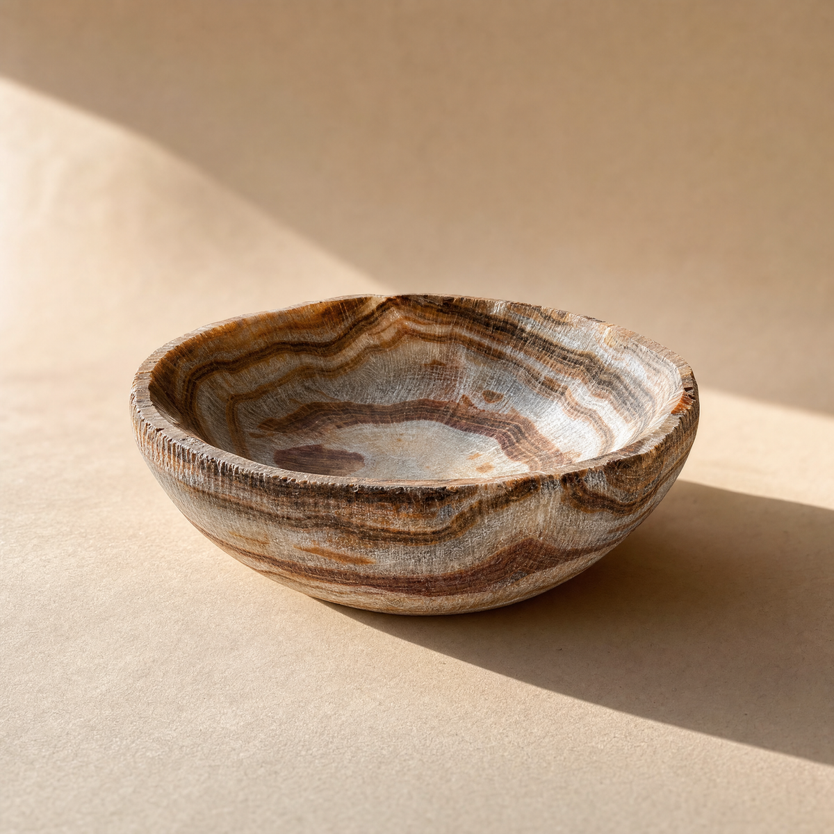 Onyx stone bowl styled on a wood table with pinecones, showing rust and cocoa striations in warm light.