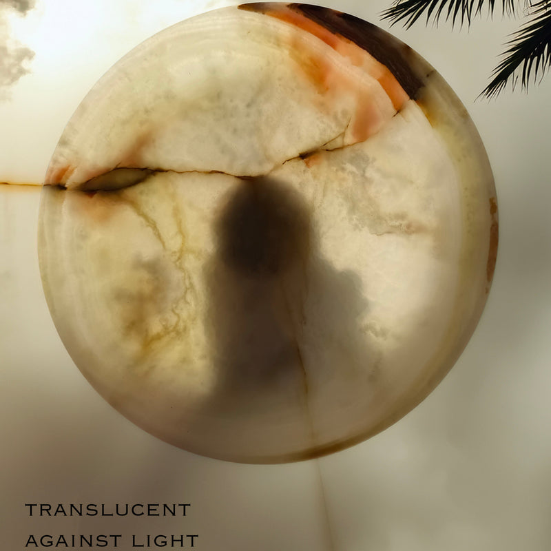 Translucent sphere with a dark spot against a light background, palm leaves visible. Top view of natural onyx pedestal bowl featuring creamy ivory stone with soft amber, green, and blush mineral veining