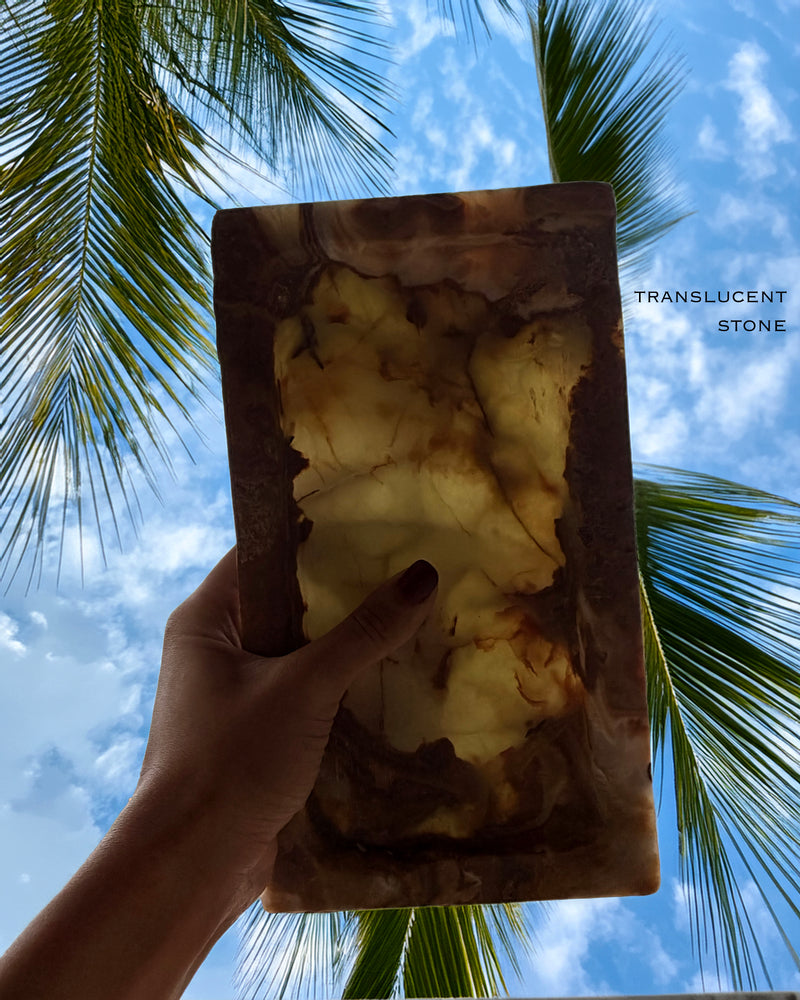 Hand holding a translucent stone with palm trees and blue sky in the background Rectangular natural onyx catchall tray with layered amber, ivory, and caramel veining photographed at an angle