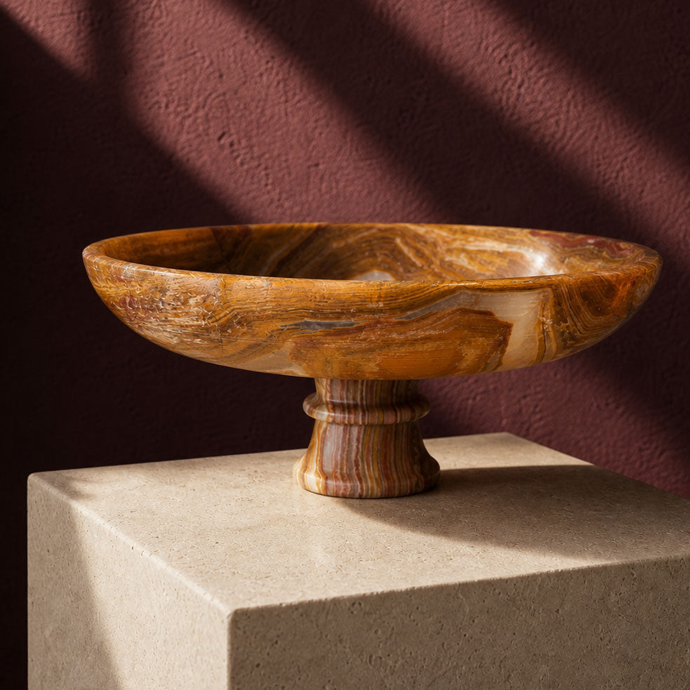 A carved onyx pedestal bowl with swirling natural brown and tan grain patterns, displayed on a light stone block against a dark reddish-brown wall with dramatic diagonal shadows.