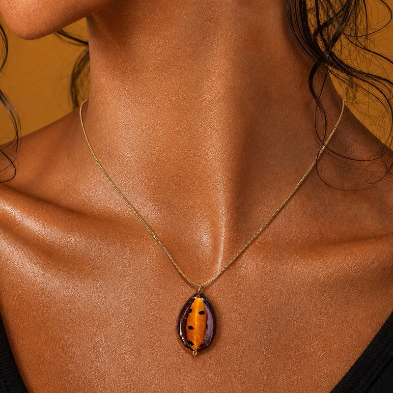 A close-up of a model's neck and chest wearing a delicate gold chain necklace with a teardrop-shaped pendant made from a polished brown and orange beetle shell.
