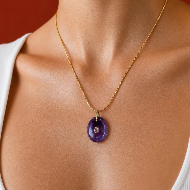 A gold chain necklace with a polished oval purple pendant, worn by a model in a white ribbed tank top against a warm red background.