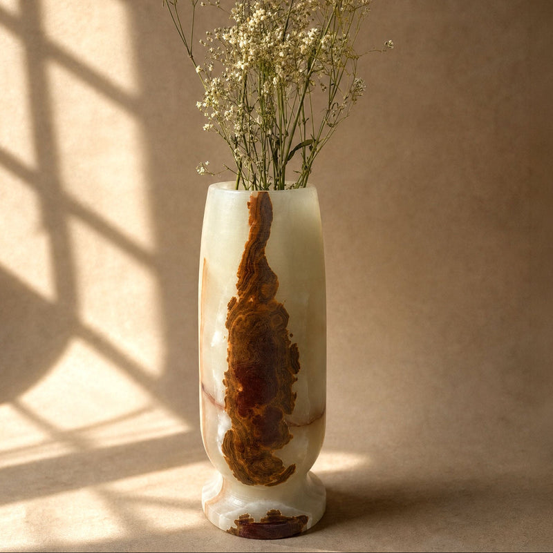 A tall, cylindrical carved stone vase with a natural brown and cream marbled pattern, filled with dried white baby's breath flowers, set against a warm beige background with soft window shadow patterns.