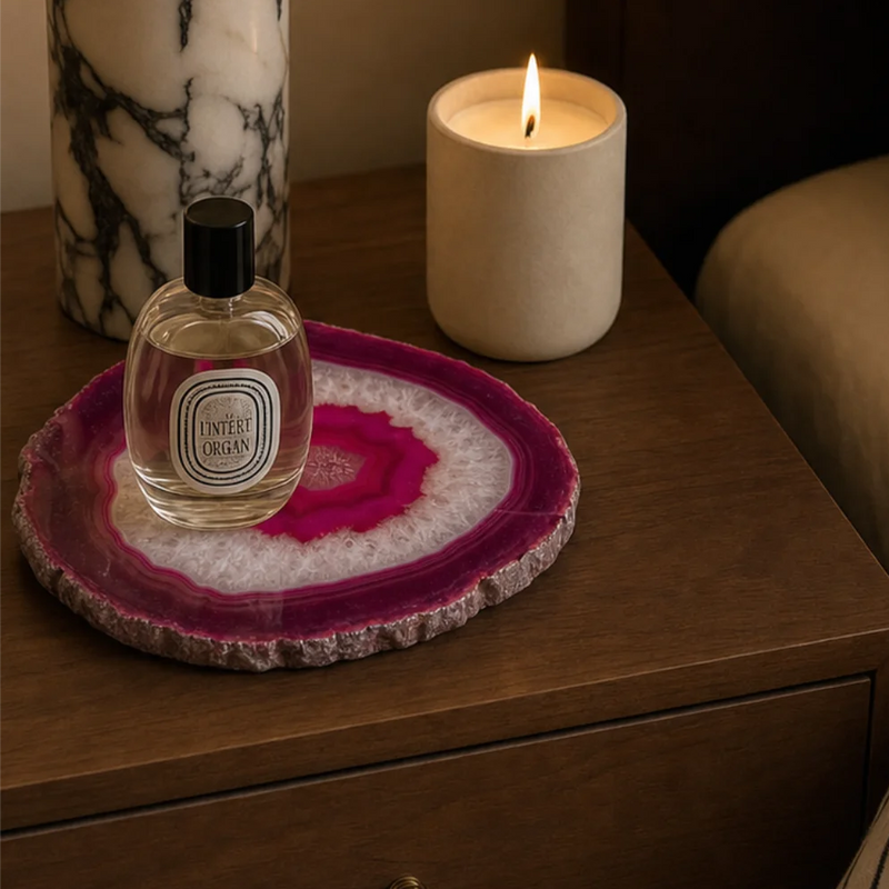A lit candle in a cream ceramic holder sits on a wooden bedside table next to a Diptyque perfume bottle and a pink agate coaster, with a marble lamp base and a bed with striped bedding in the background.