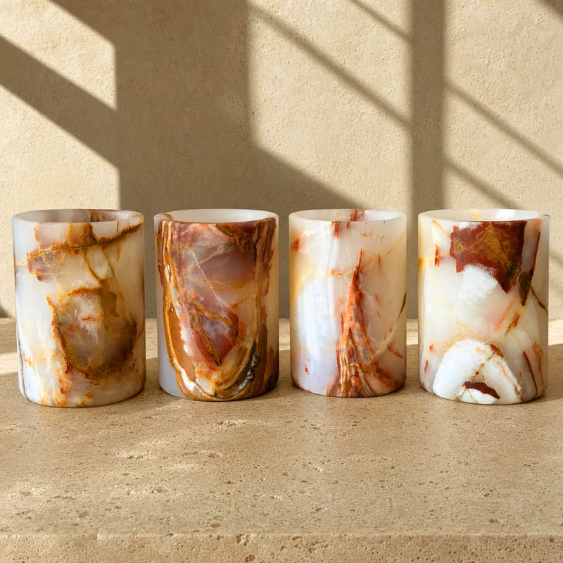Four cylindrical onyx stone vessels with marbled onyx-like patterns in white, cream, and warm brown tones, arranged side by side on a textured stone surface against a beige wall with soft window-shadow lighting.
Four one-of-one blush and rust Balochistan onyx cylinder vessels lined up together, showing the range of veining in a rare pink-toned pull.