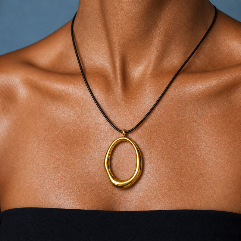 A close-up of a model's neck and chest wearing a black cord necklace with a polished gold abstract oval pendant.