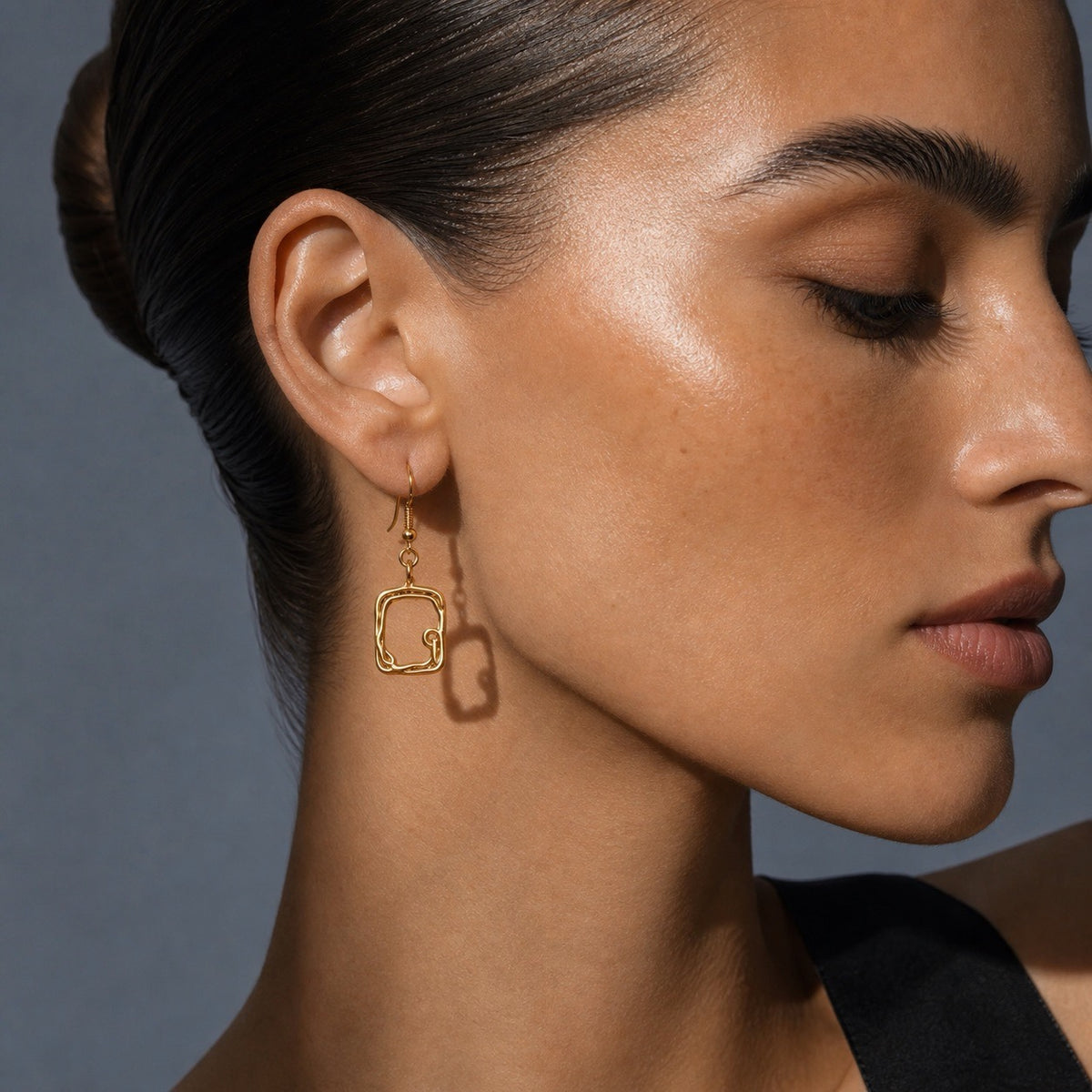 A close-up side profile of a woman wearing a delicate gold hoop earring with a small square dangling charm, set against a dark gray background.