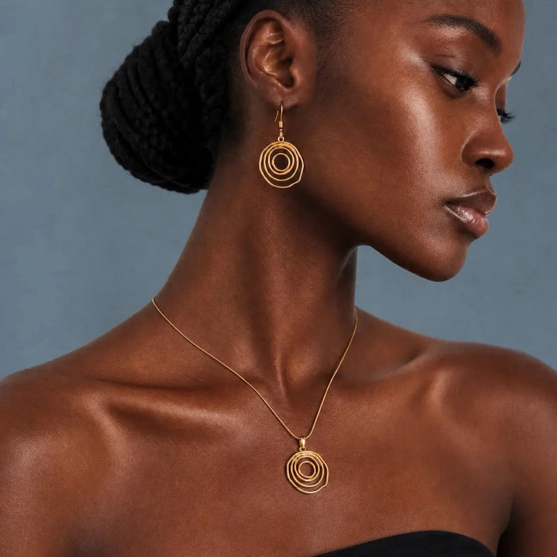 A close-up of a woman wearing a gold spiral pendant necklace and matching spiral drop earrings against a muted blue-gray background.