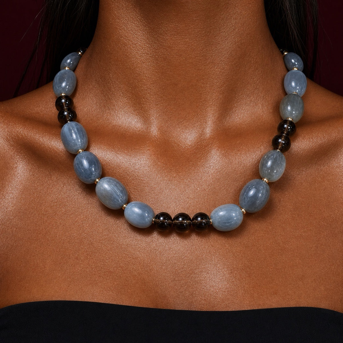 A close-up of a beaded necklace worn on a model's neck. The necklace features large, polished light blue oval beads separated by small gold spacers, with a section of smaller dark brown and black beads.