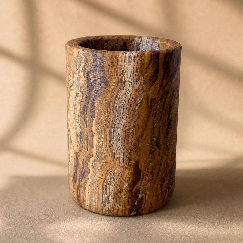 A cylindrical onyx cup or vase made from polished stone with rich brown, gold, and black wavy grain patterns, set against a warm beige background with soft window shadow overlays.