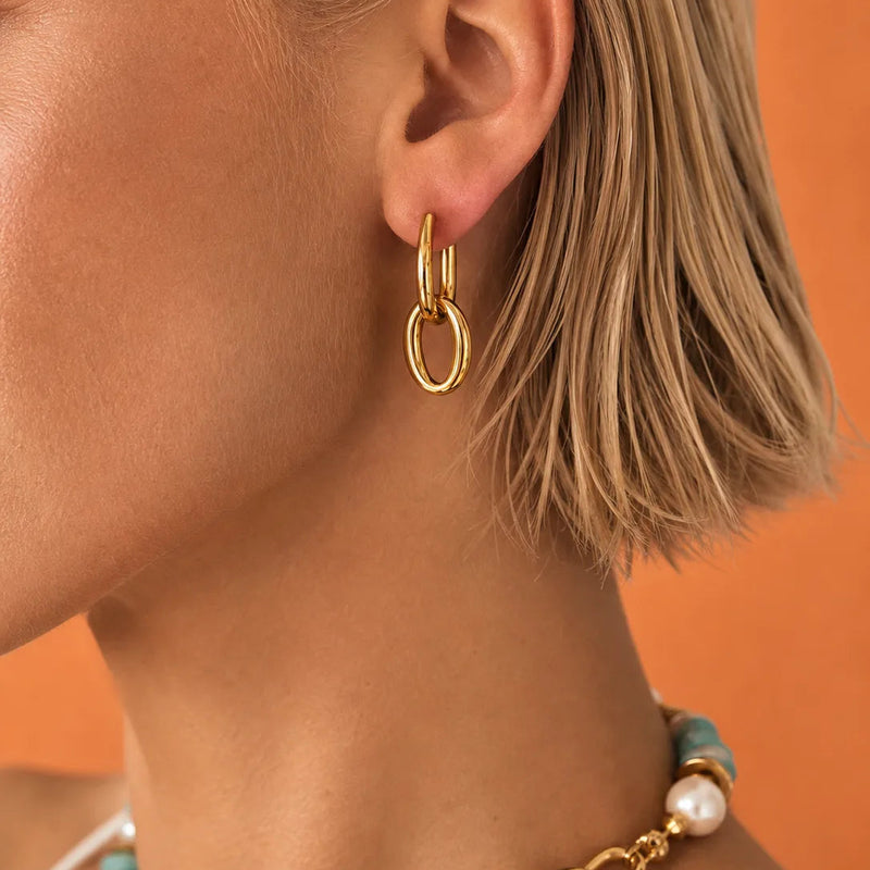 A close-up of a model's ear wearing a pair of gold hoop earrings, with a matching gold chain necklace partially visible. The model has short blonde hair and is set against a solid orange background.