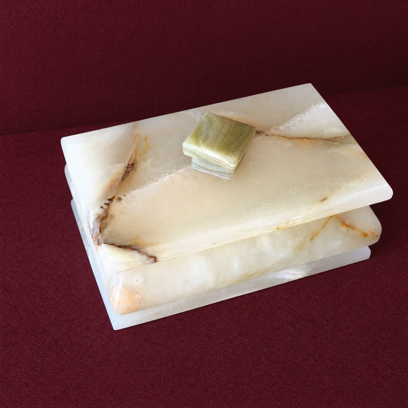 A rectangular cream-colored onyx stone box with a matching lid, featuring natural brown and beige veining, with a small green stone accent on top, set against a dark red background.