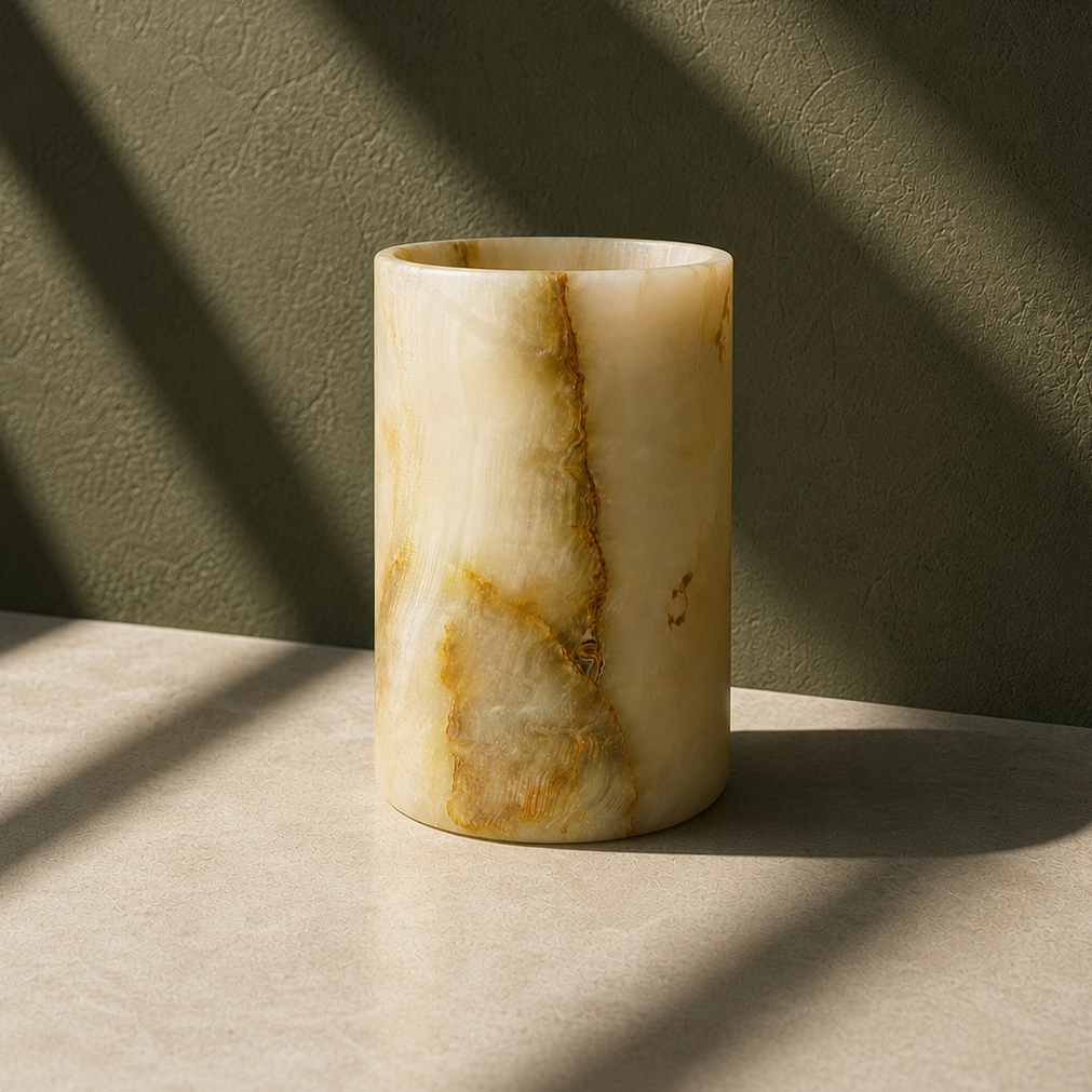A cylindrical stone vase made of cream and tan marbled onyx or alabaster, standing on a light stone surface with dramatic diagonal shadows cast across a textured olive-green wall.