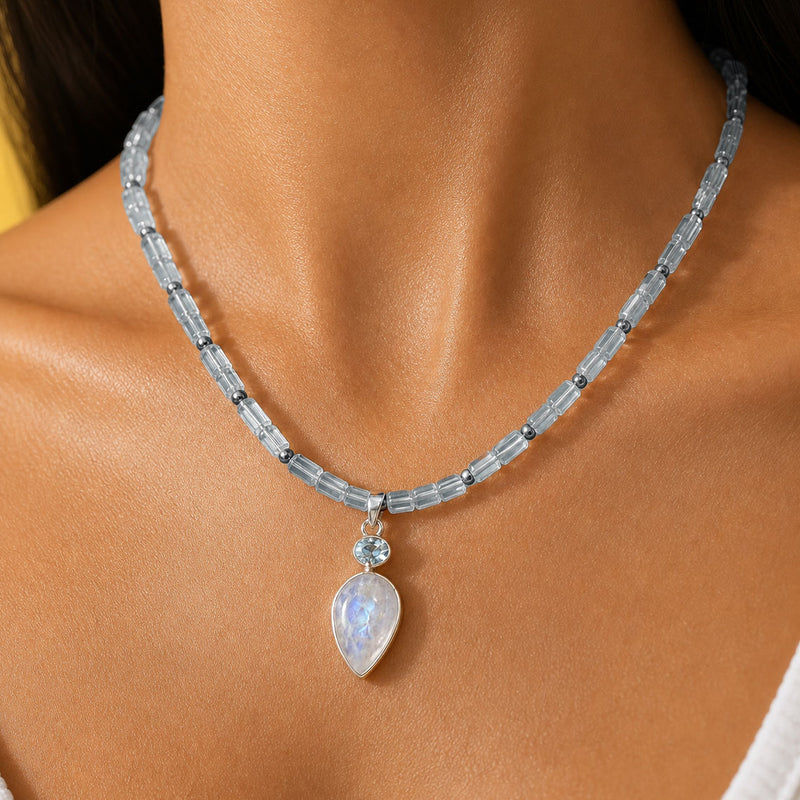 A close-up of a woman's neck and chest wearing a beaded necklace with a teardrop-shaped iridescent moonstone topaz pendant. The necklace features light blue faceted topaz beads separated by small dark accents, with a silver-toned clasp and a small clear stone above the pendant.