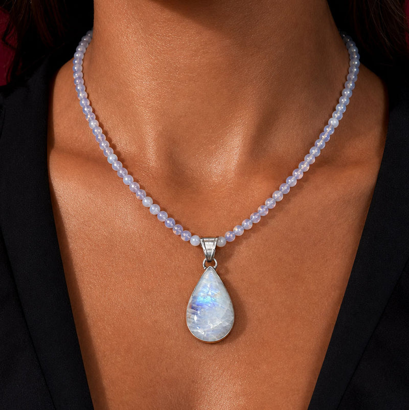 A close-up of a woman's neck and chest wearing a beaded necklace with a teardrop-shaped iridescent pendant. The necklace features pale lavender-beige beads and a silver-toned bail holding a white teardrop pendant with blue and iridescent sheen.