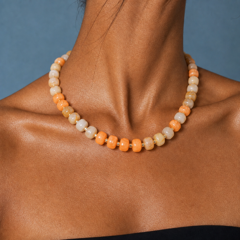 A beaded necklace with alternating orange and cream-colored round beads, shown worn on a model's neck against a blue-gray background.