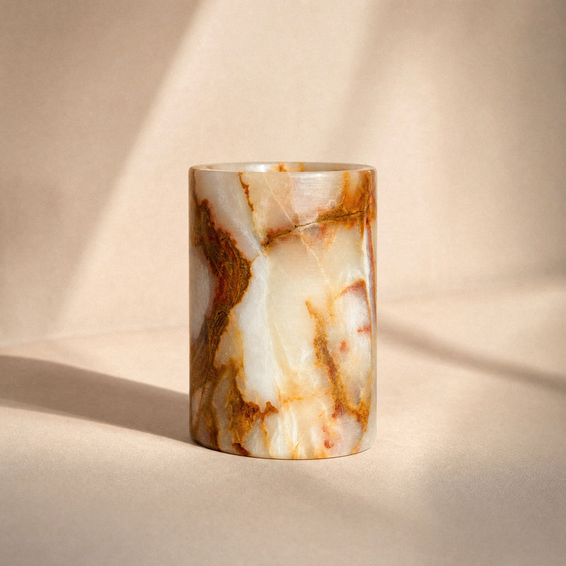 Translucent cream onyx vessel with amber and rust veining holding two bamboo toothbrushes on a marble bathroom vanity