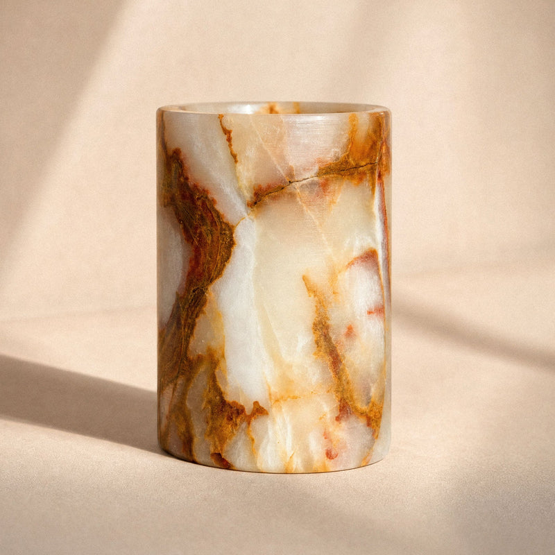Translucent cream onyx vessel with amber and rust veining holding two bamboo toothbrushes on a marble bathroom vanity