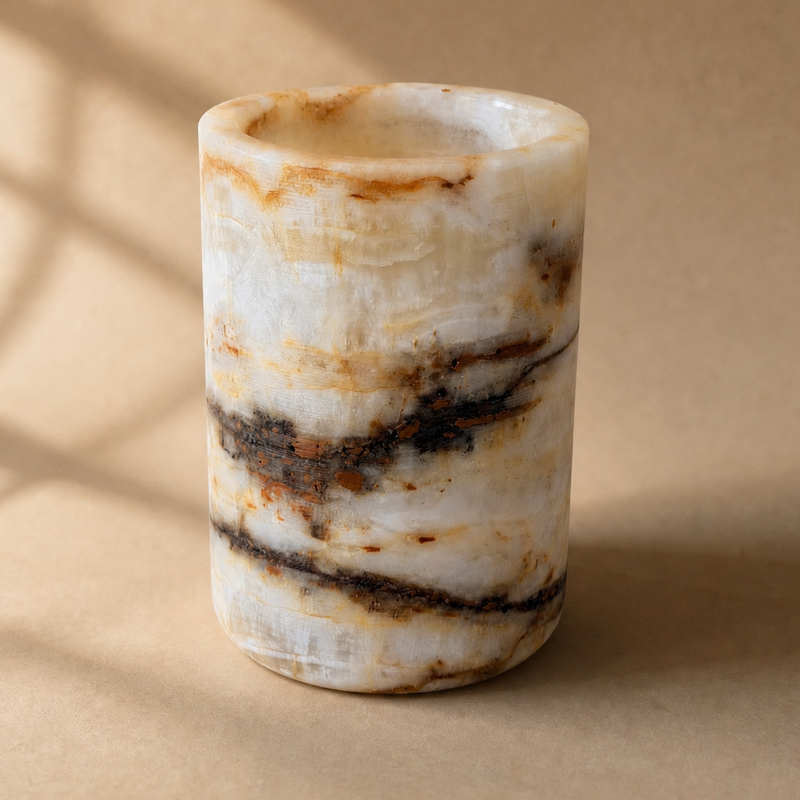 A small cylindrical stone tumbler or candle holder made of marbled white, beige, and brown stone, photographed on a warm beige surface with soft window-shadow light.