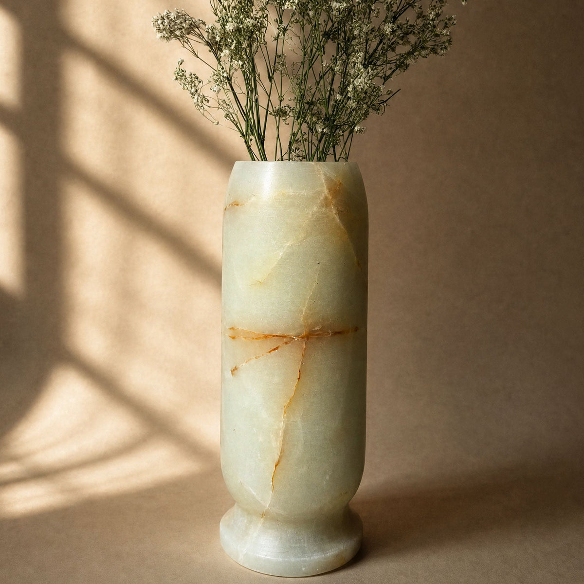 A cylindrical pale green marble vase with natural veining, filled with dried baby's breath flowers, set against a warm beige background with soft window shadow patterns.