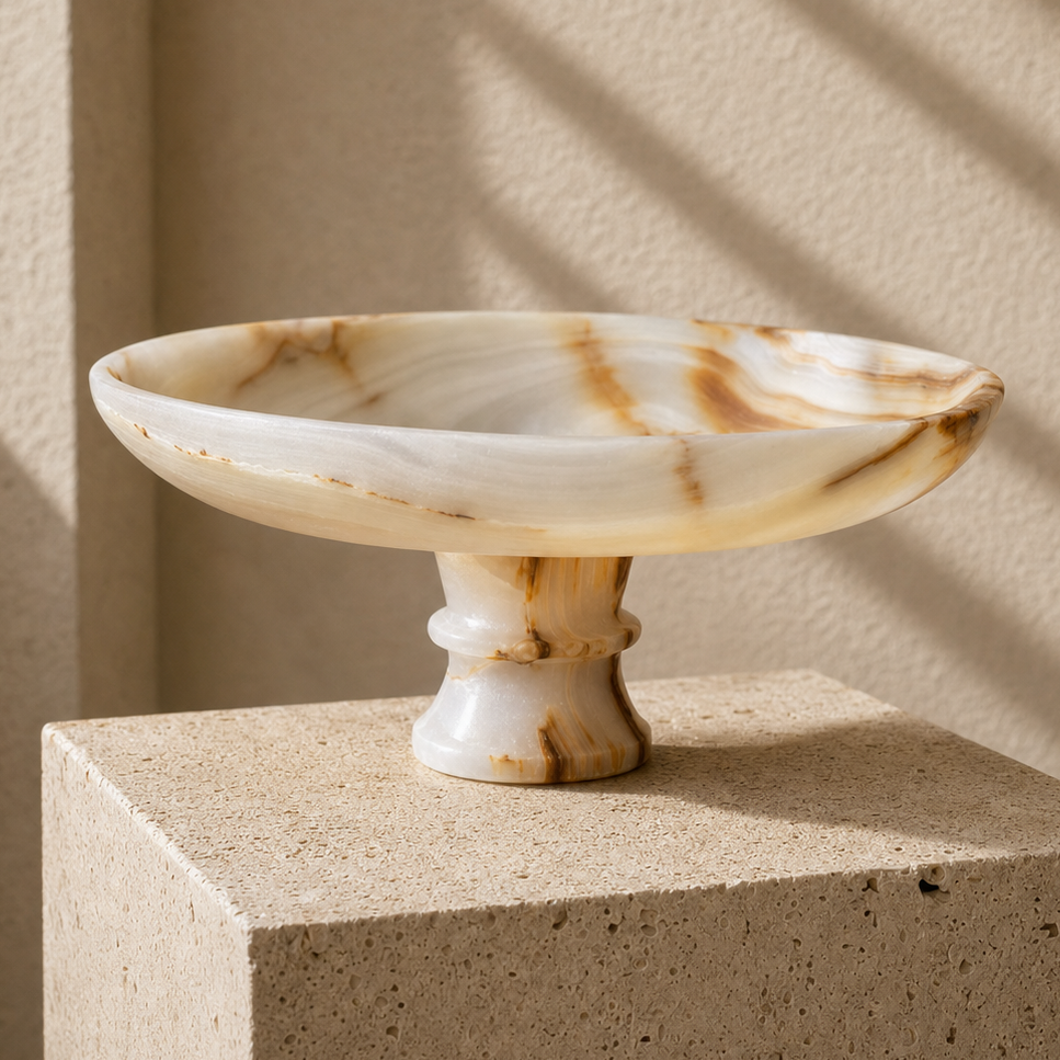 A round onyx pedestal bowl made of natural marbled stone in cream, beige, and brown tones, displayed on a textured stone block against a beige wall with soft diagonal shadows.