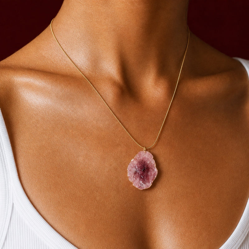 A close-up of a model's neck and chest wearing a delicate gold chain necklace with a raw, irregular pink geode-style pendant.