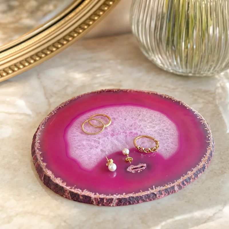 A round agate slice jewelry tray with a vibrant magenta and pink patterned center and a natural brown textured edge, styled on a marble surface with gold rings and pearl earrings.