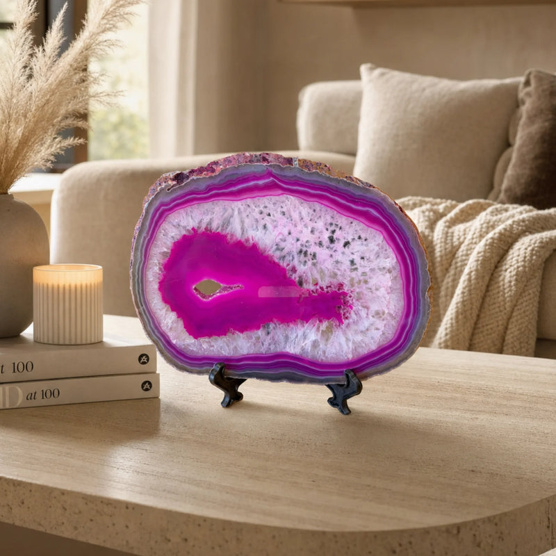 A polished agate geode slice with vivid magenta, purple, and white banding, displayed on a small black stand on a light wood table in a cozy living room setting.