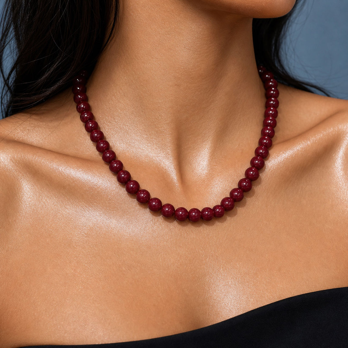 A close-up of a model's neck and shoulders wearing a dark red beaded necklace against a soft blue-gray background.