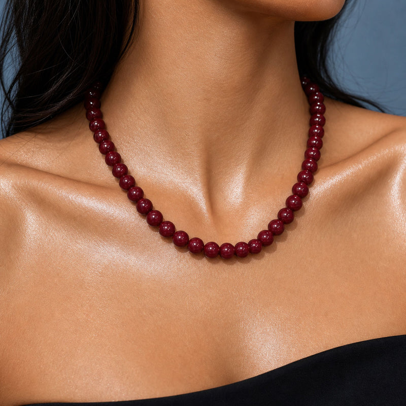A close-up of a model's neck and shoulders wearing a dark red beaded necklace against a soft blue-gray background.