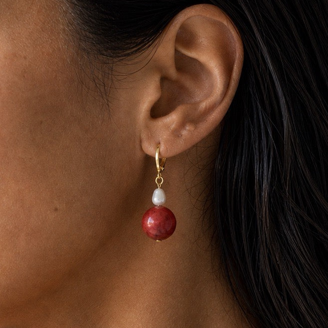 A close-up of a model's ear wearing a gold huggie hoop earring with a small white freshwater pearl drop and a round red coral stone pendant.