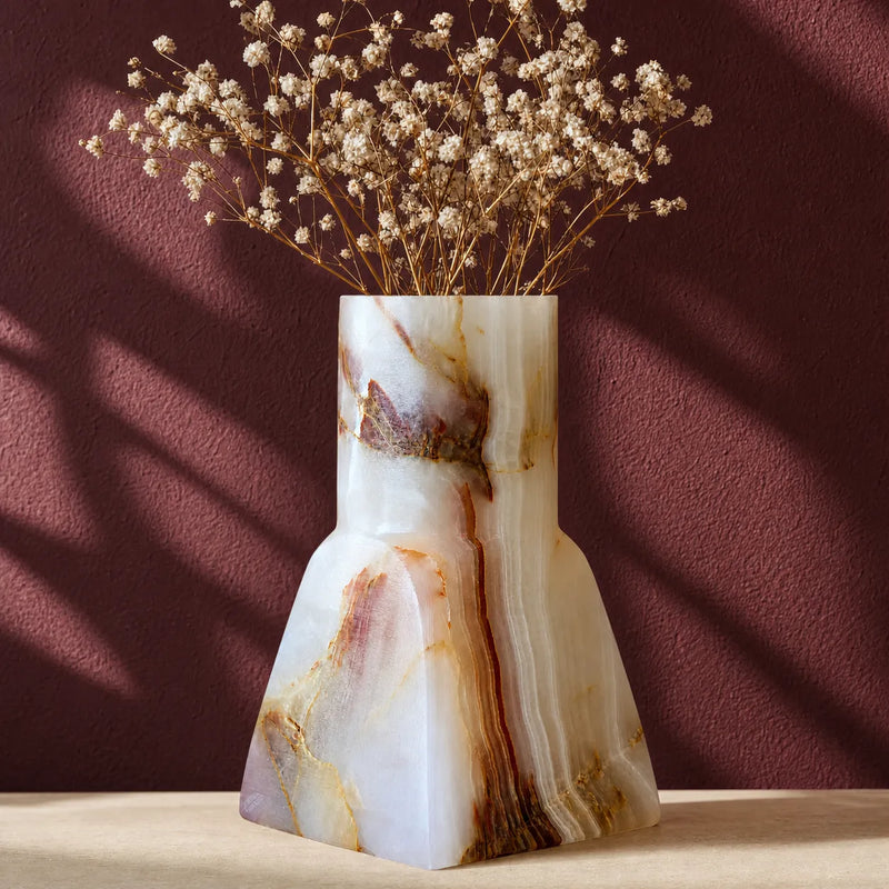 A sculptural onyx-style vase with a tapered silhouette, featuring natural cream, white, and warm brown marbling. The vase is filled with dried baby's breath flowers and is set against a textured dark burgundy wall with soft diagonal shadows.