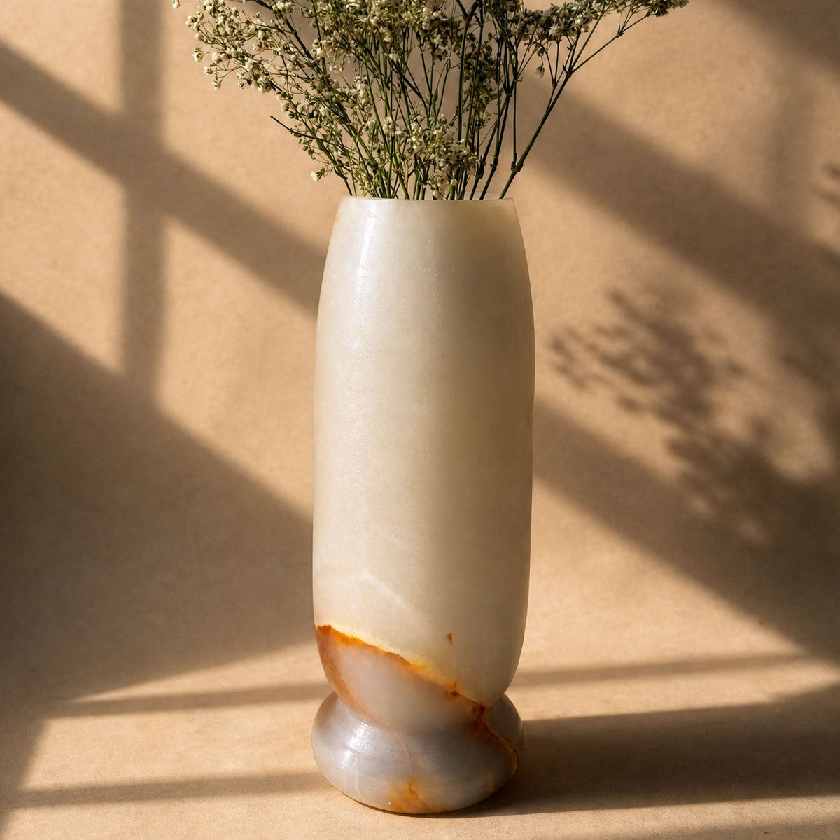 A tall, cylindrical off-white stone vase with a natural brown and gray marbled base, holding dried baby's breath flowers, set against a warm beige background with soft window shadow patterns.