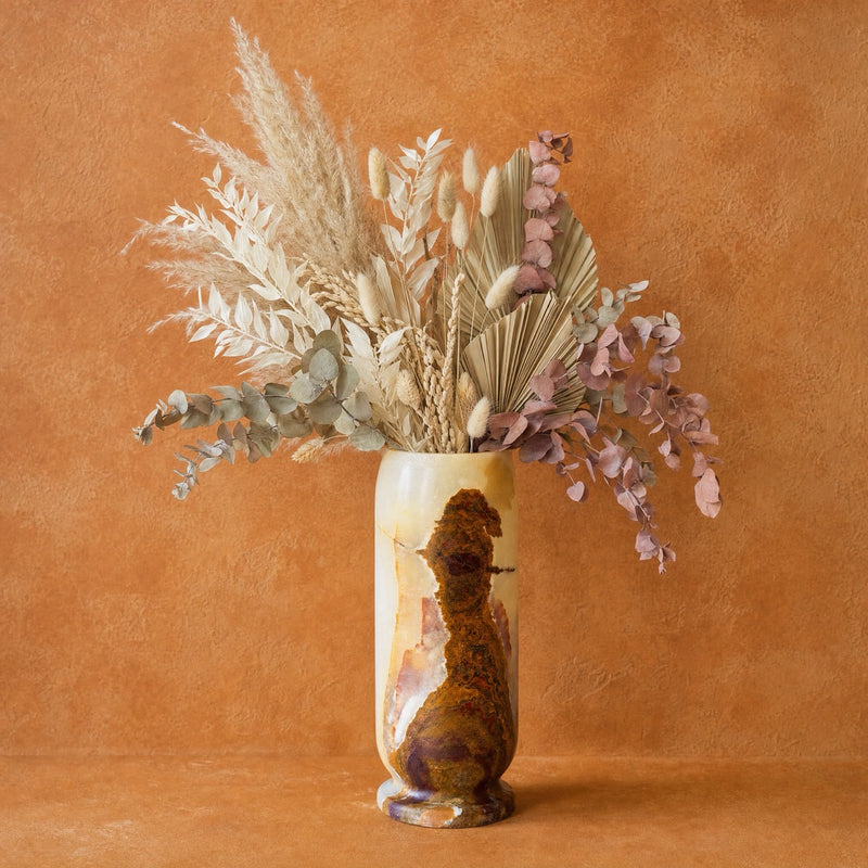 A decorative onyx vase with a glossy brown and gold abstract design, filled with a dried floral arrangement of beige pampas grass, eucalyptus, and dusty pink foliage, set against a warm textured terracotta background.
