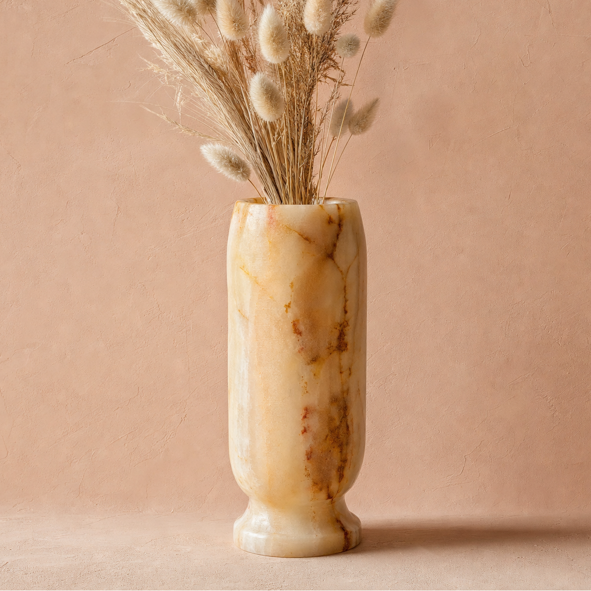 A cylindrical onyx marble vase with a warm cream and tan base and brown veining, filled with dried bunny tail grass, set against a textured dusty pink background.