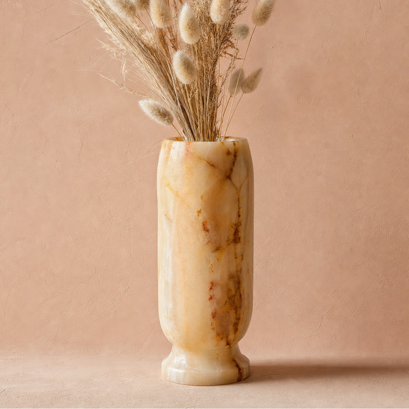 A cylindrical onyx marble vase with a warm cream and tan base and brown veining, filled with dried bunny tail grass, set against a textured dusty pink background.