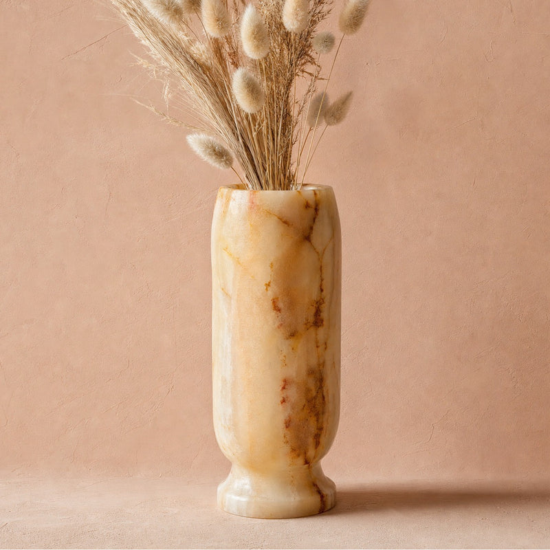 A tall cylindrical onyx vase in warm cream and tan tones with natural brown veining, filled with dried bunny tail grass, set against a soft dusty pink background.
