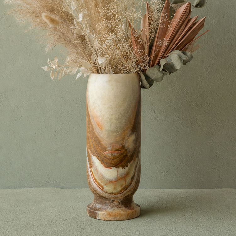 A tall cylindrical onyx vase made of marbled stone in cream, tan, and brown tones, filled with dried grasses and botanical stems, set against a muted sage green wall and surface.