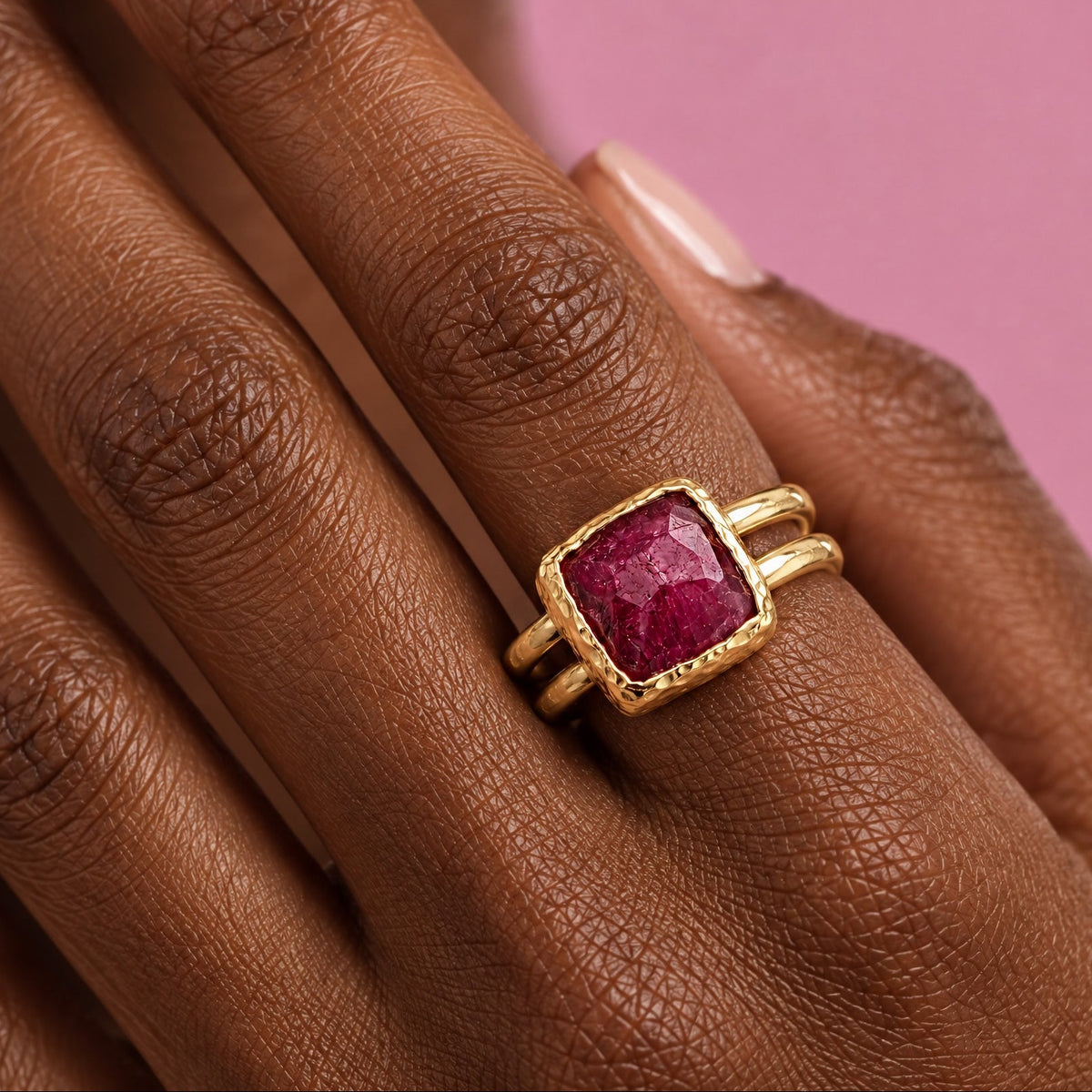 Close view of a hand wearing an adjustable square ruby ring, the faceted raspberry red stone in a hammered 22k gold-filled bezel on a split double band, against pink