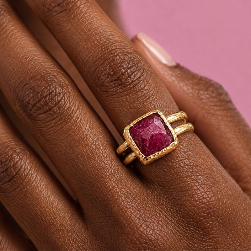 Close view of a hand wearing an adjustable square ruby ring, the faceted raspberry red stone in a hammered 22k gold-filled bezel on a split double band, against pink