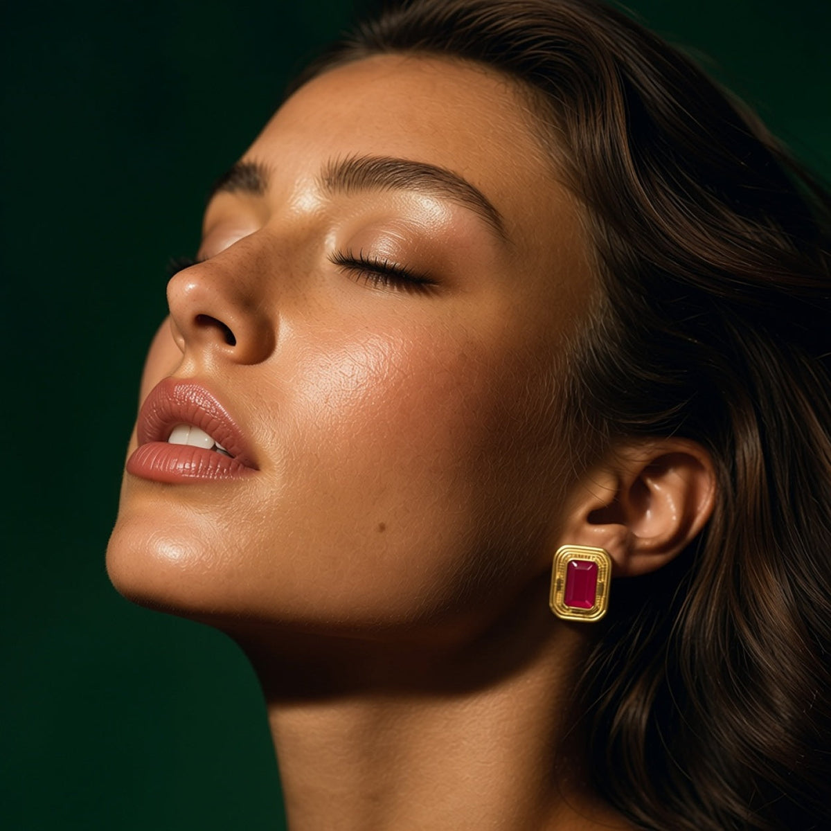 Model with her eyes closed wearing the berry cat's eye stud earring in its stepped gold frame, against a deep green background.