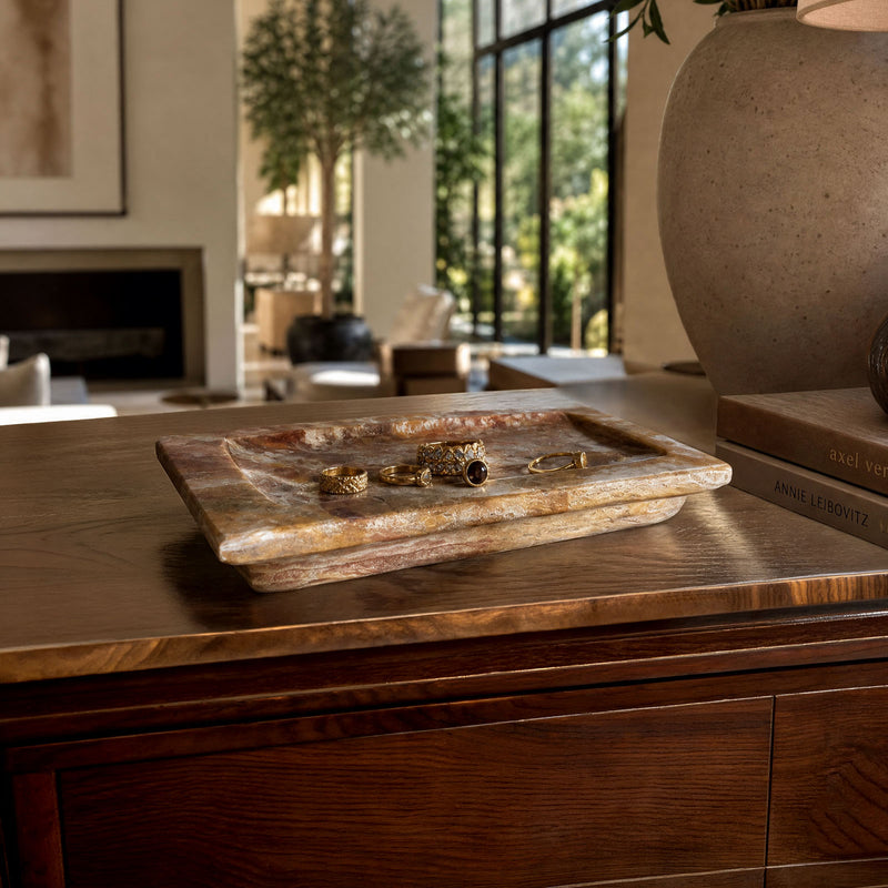 Rustic onyx stone tray holding gold rings and jewelry on a walnut dresser in a sunlit modern living room with olive tree and stone vase