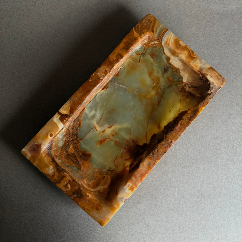 Rectangular stone with marbled pattern on a gray background Hand-carved raw onyx vanity tray in cream, honey, and warm brown tones styled with perfume, watch, and hand cream on a neutral surface