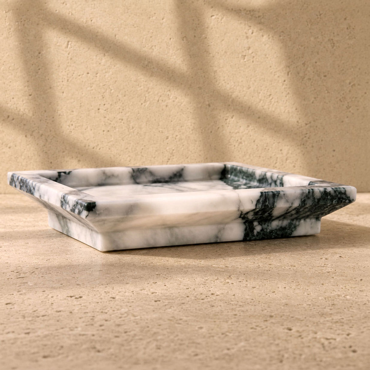 A rectangular marble tray with a white base and dark green veining, set on a textured beige stone surface with soft window shadow patterns. White Calacatta Vagli marble catchall tray with deep green and charcoal veining, raised rim on a plinth base, on a travertine surface