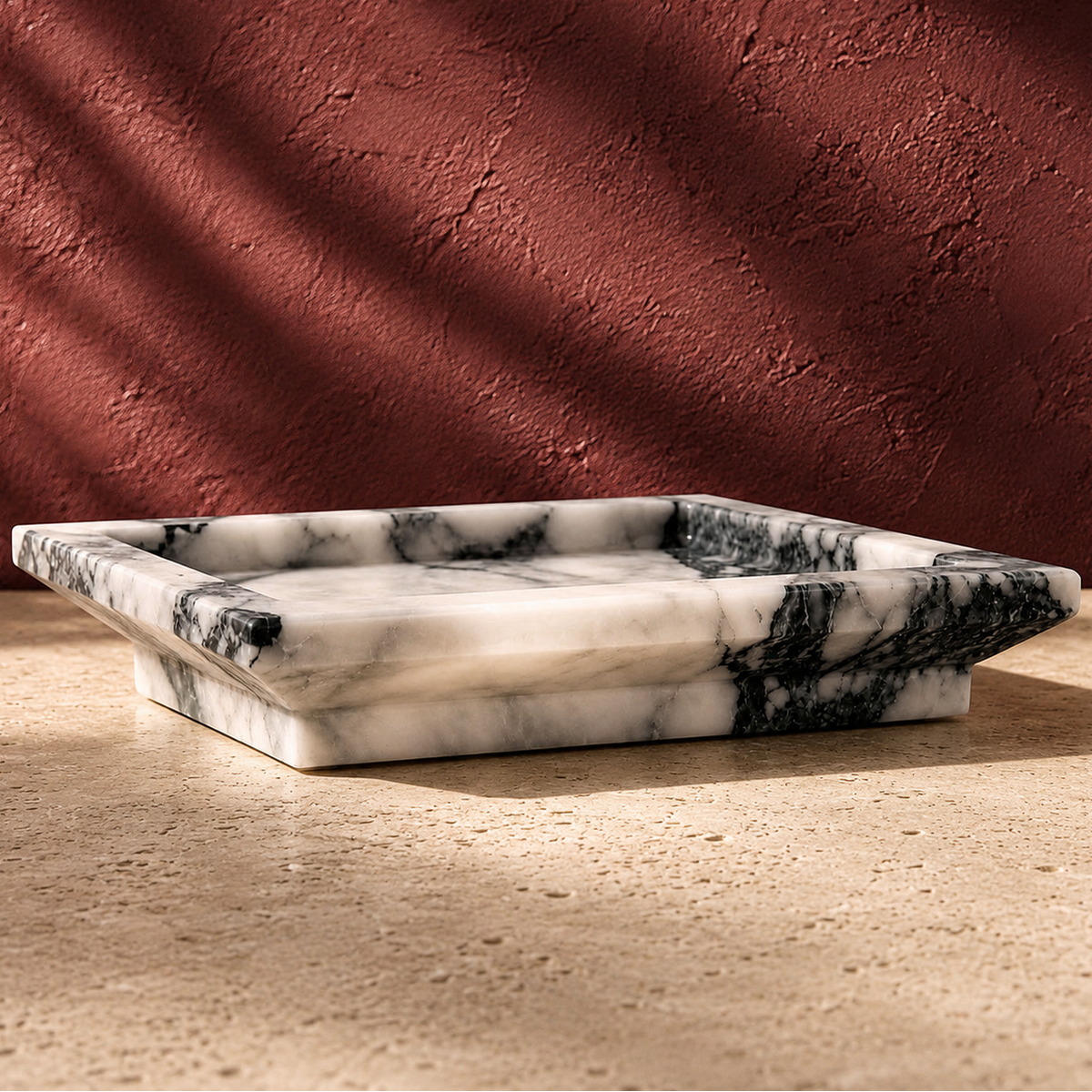 A rectangular white calacatta viola tray with black and gray veining, set on a textured beige stone surface against a terracotta wall with dramatic leaf shadows.