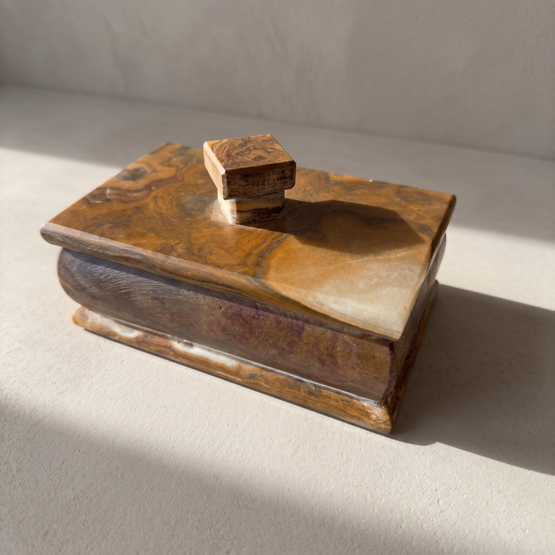 Onyx keepsake box with a fitted lid, three-quarter front view, caramel and honey swirls on the lid ending in a translucent cream corner, walnut-brown bulged body with a plum blush, pale plaster surface in raking sun