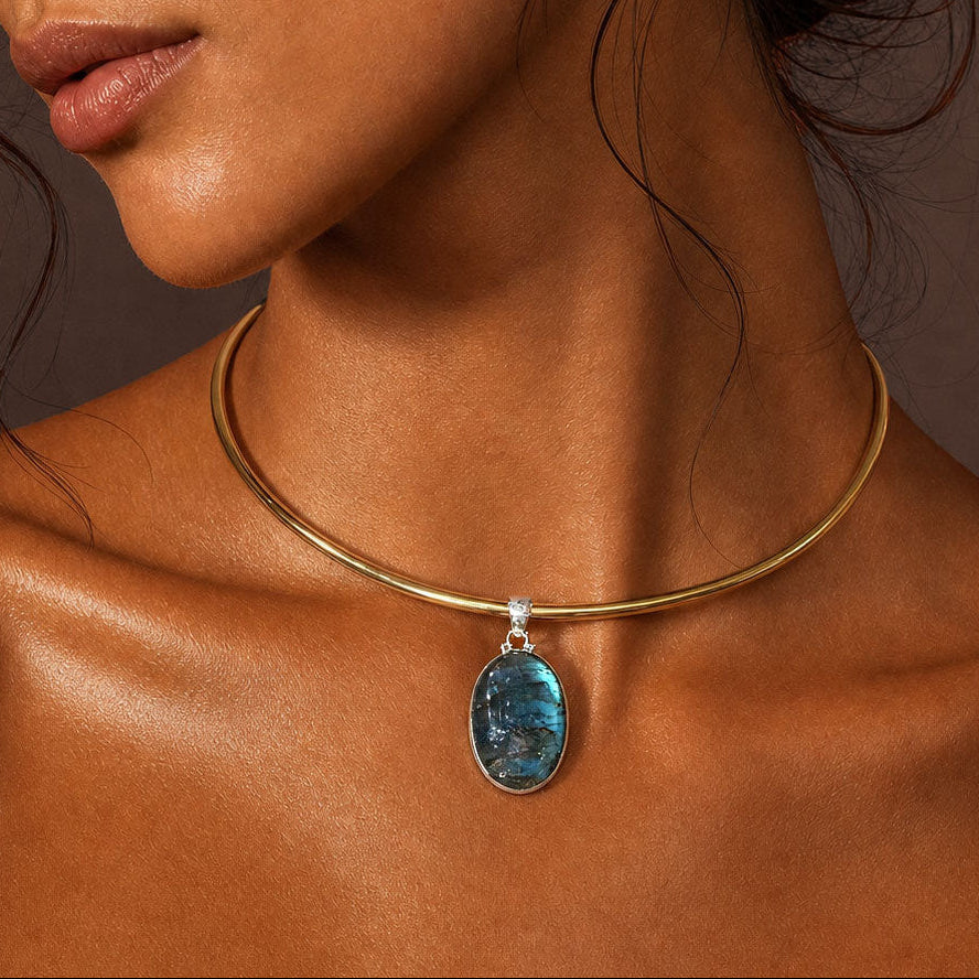 A close-up of a model wearing a delicate gold collar necklace with a dangling oval blue labradorite gemstone pendant.