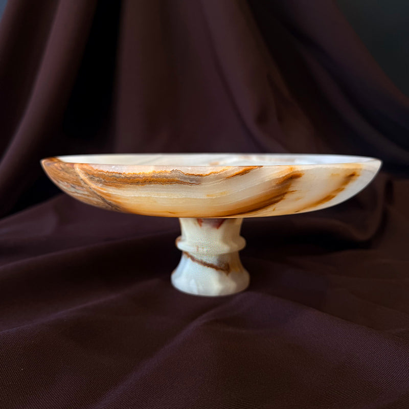 Three-quarter view of the hand-carved onyx pedestal bowl, amber and cream stripes across the outer wall of the shallow basin